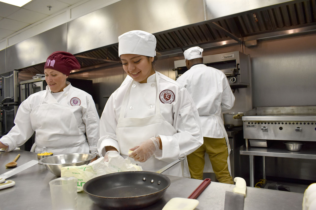 Culinary arts students stand in front of metal table and measure out ingredients